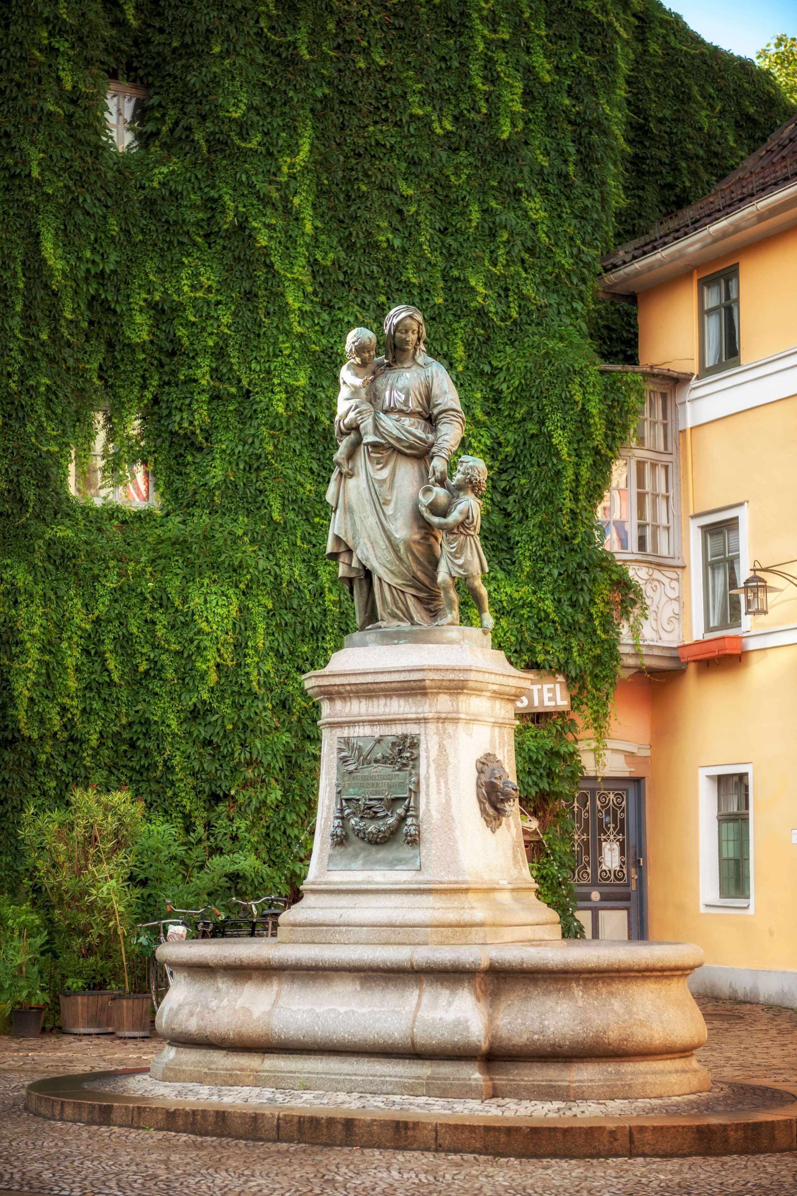 A statue of a woman holding a child, set on a pedestal with a lion emblem, in front of a building with a 'TEL' sign.