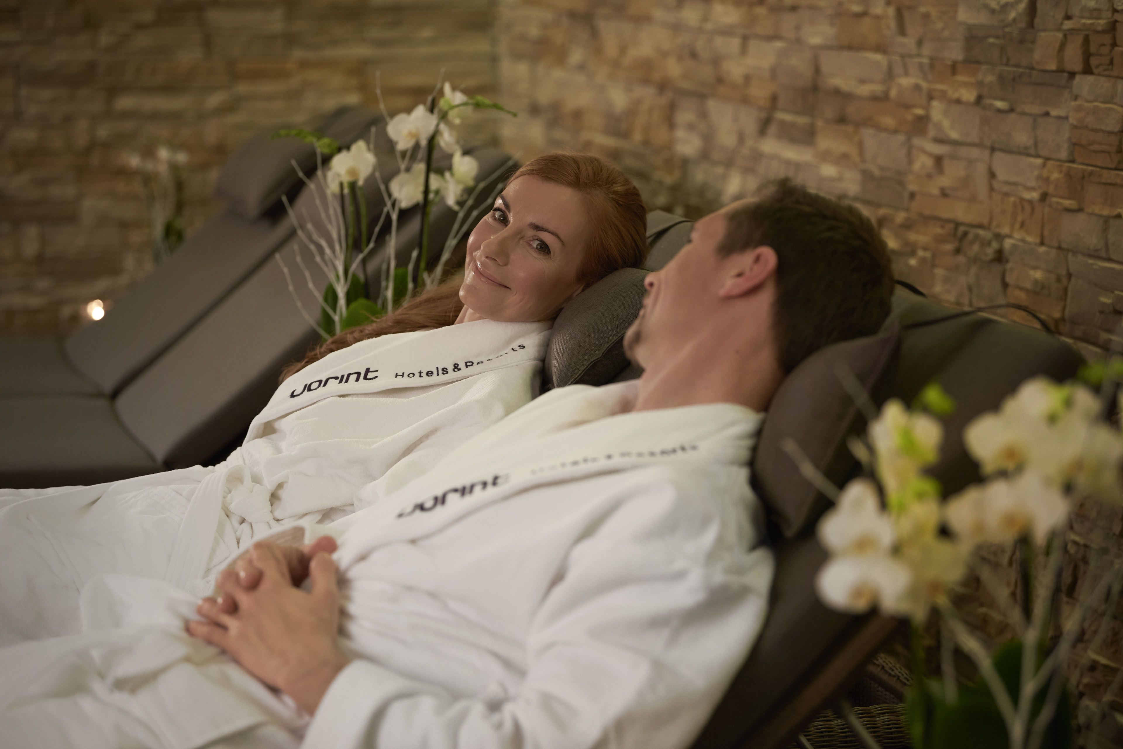 Two people lying on reclining chairs in a spa setting with a stone wall in the background.