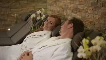 Two people lying on reclining chairs in a spa setting with a stone wall in the background.