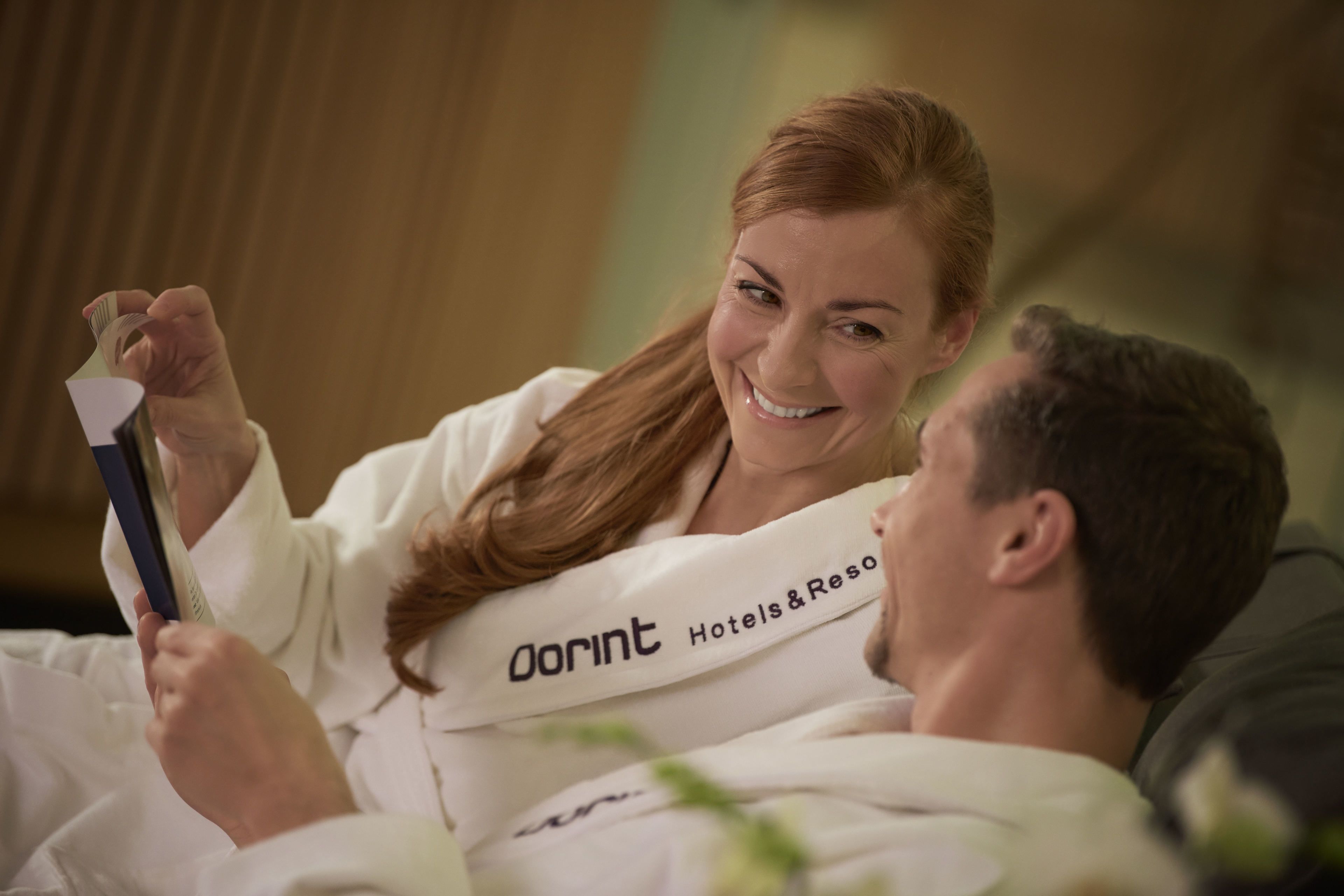 A person in a white robe with 'DORINT Hotels & Resorts' on it, lying in bed with a smartphone.