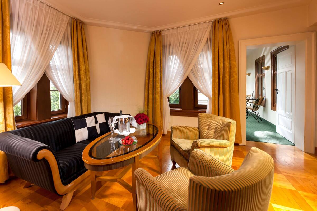 A room with a sofa, armchairs, a glass coffee table, and a lamp. There are curtains, a window, and a door visible.