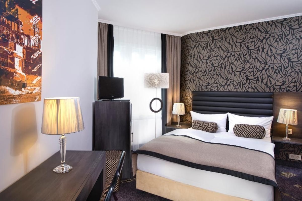 Habitació d'hotel amb un llit, una televisió, un escriptori amb una làmpada i una paret amb un disseny decoratiu.