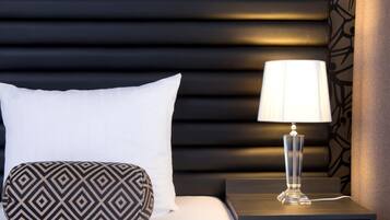 Una cama bien hecha con un almohadón blanco y otro estampado, una mesita de noche con una lámpara, y una pared con un diseño geométrico.