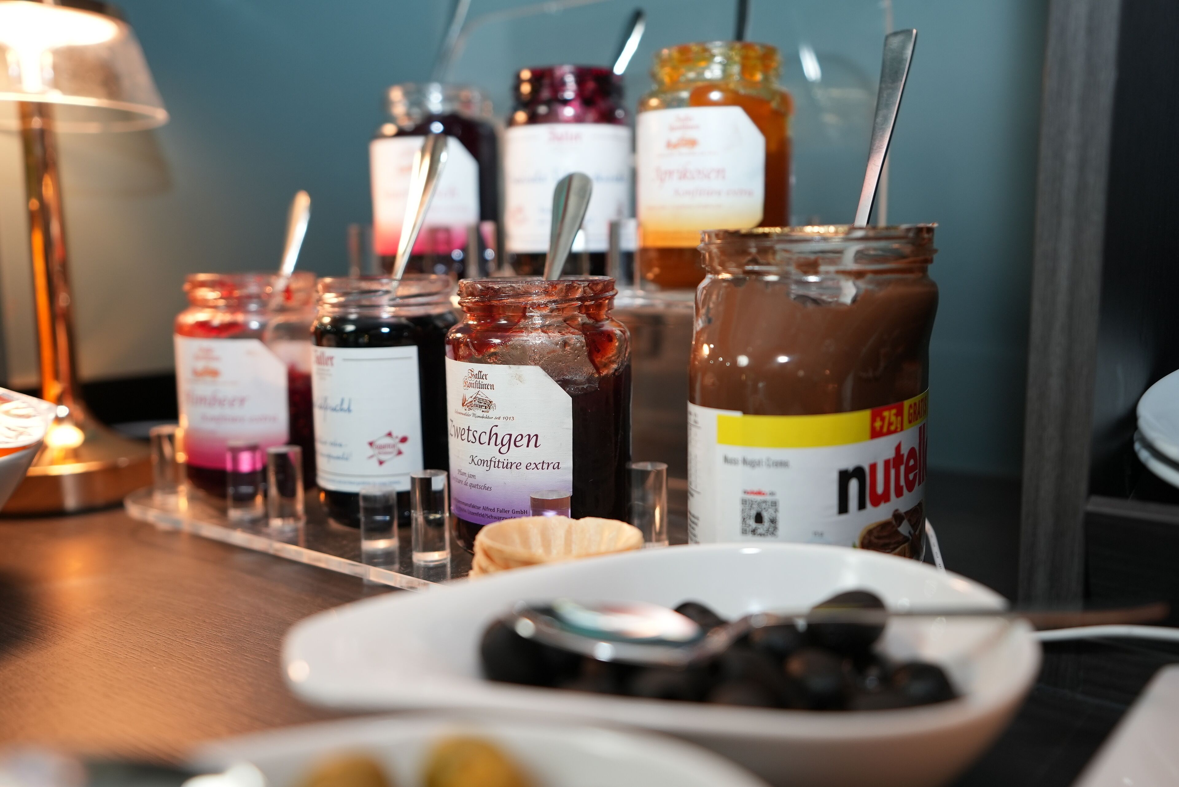 A table with various jars of spreads and condiments, including Nutella, and a plate with olives.