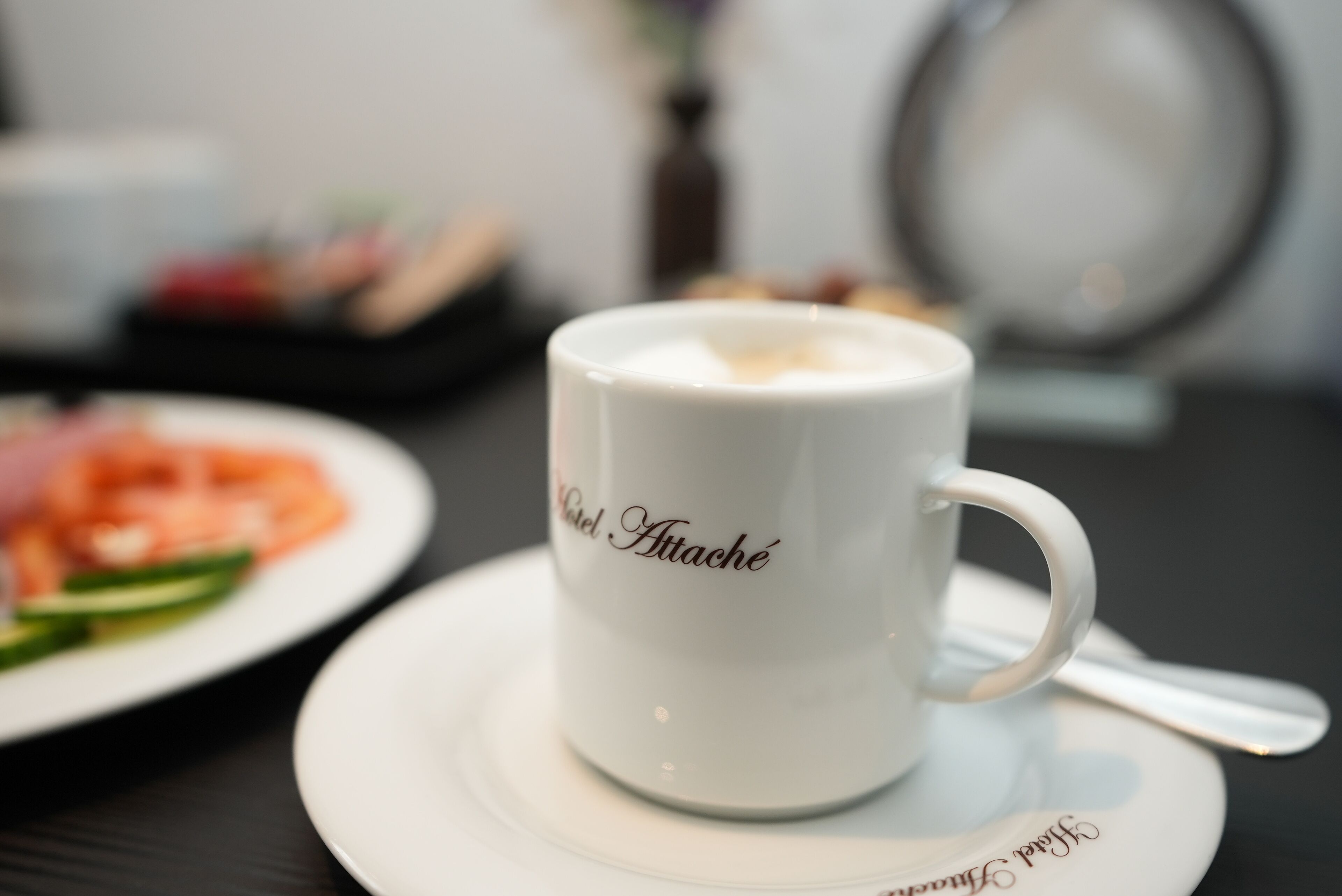 Eine weiße Kaffeetasse mit dem Aufdruck "Café Attache", platziert auf einem Untertasse mit einem Löffel daneben.