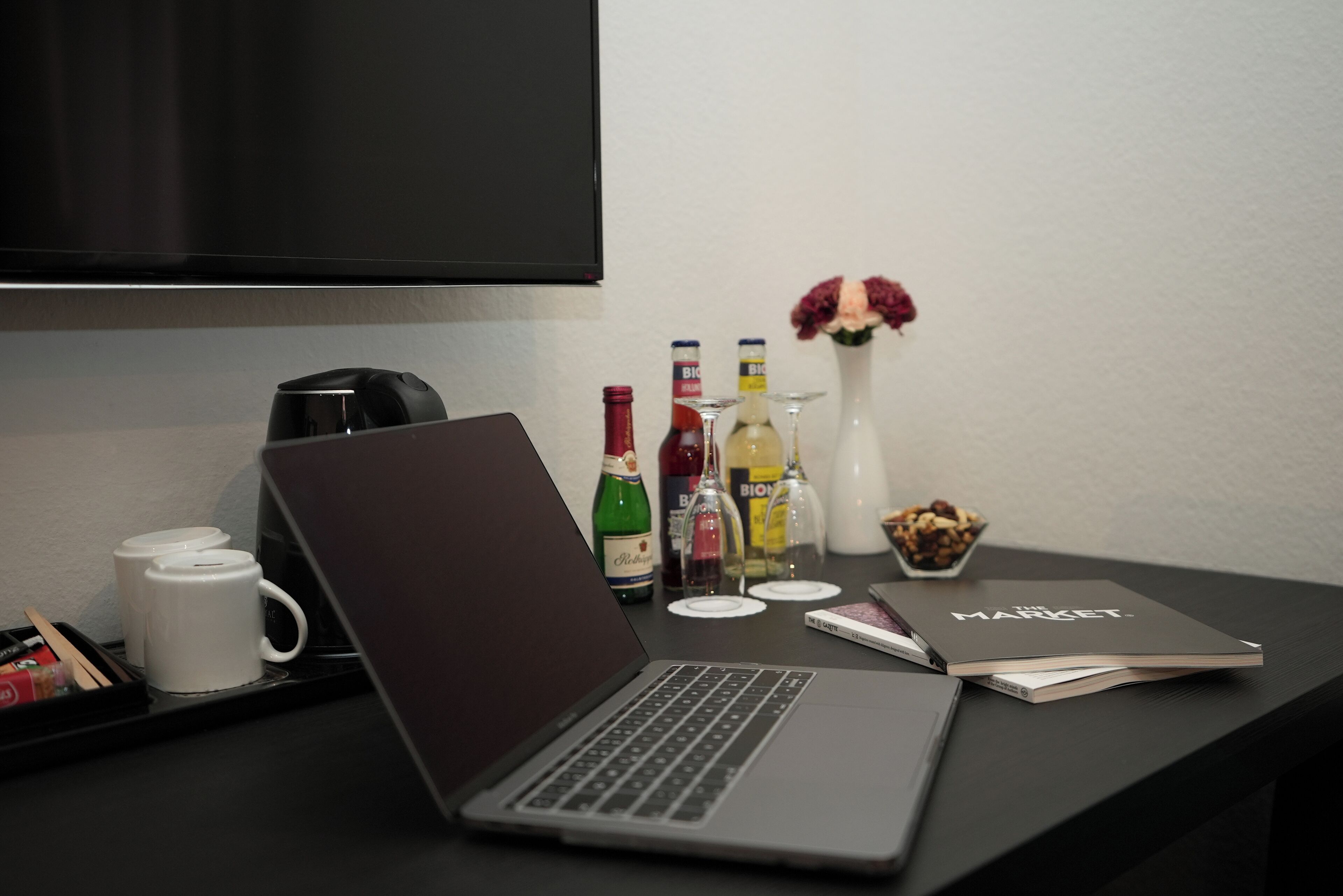 A laptop on a desk with a magazine, a coffee maker, and various bottles.