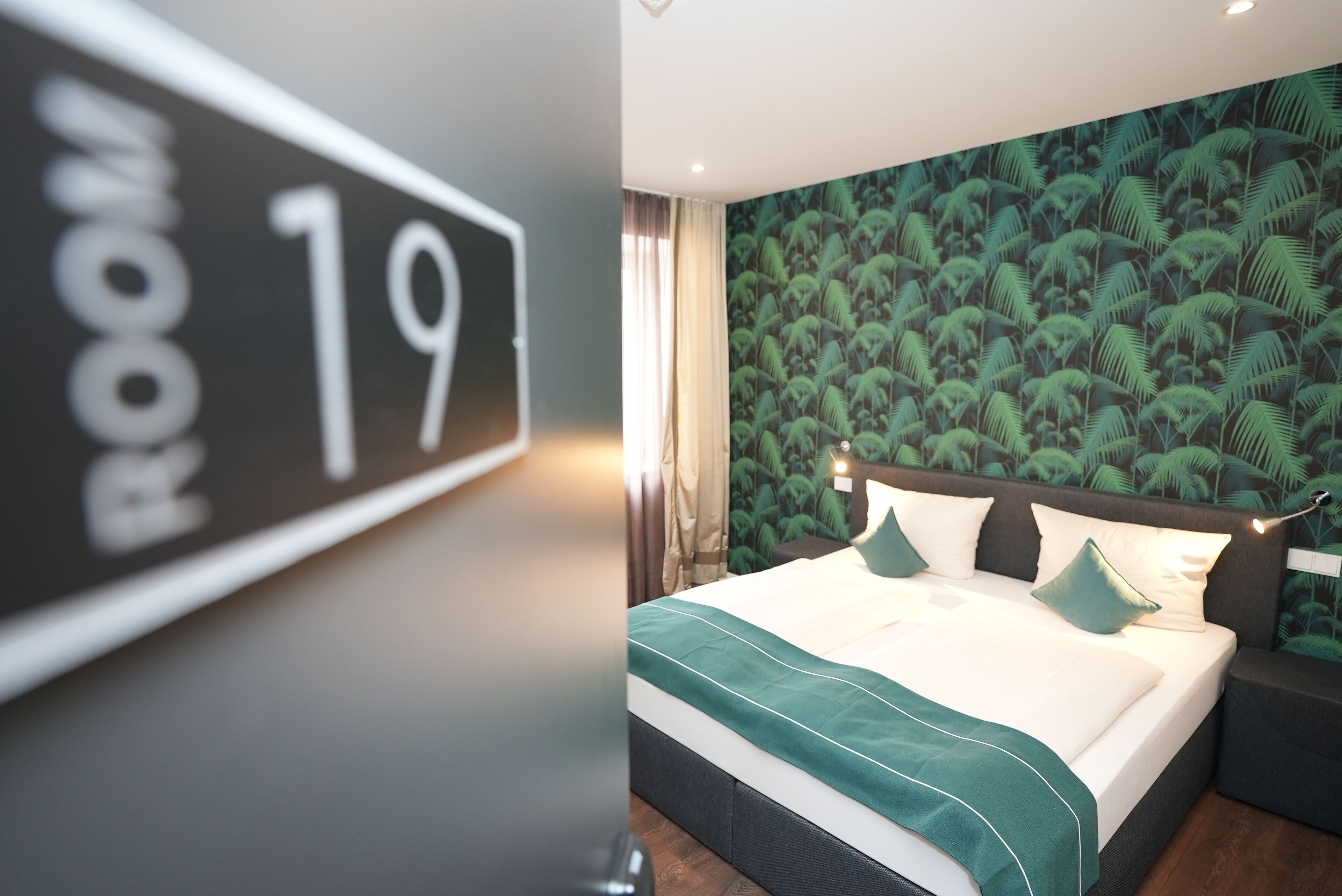A hotel room with a bed, a headboard, a wall with a tropical leaf pattern, and a sign indicating the room number 19.