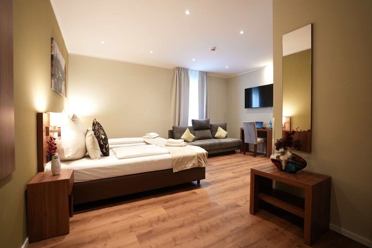 A modern hotel room with a large bed, a sofa, a TV, and a wooden floor.