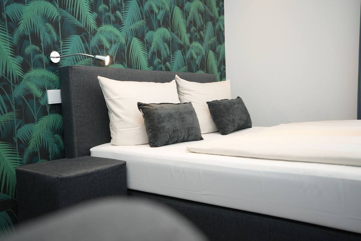 A modern bedroom with a bed, dark headboard, white bedding, and a wall with a green leaf pattern.