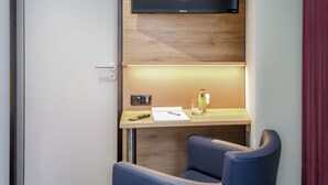 Una habitación de hotel compacta con un escritorio de madera, una silla azul, un televisor de pantalla plana y una lámpara de mesa pequeña.