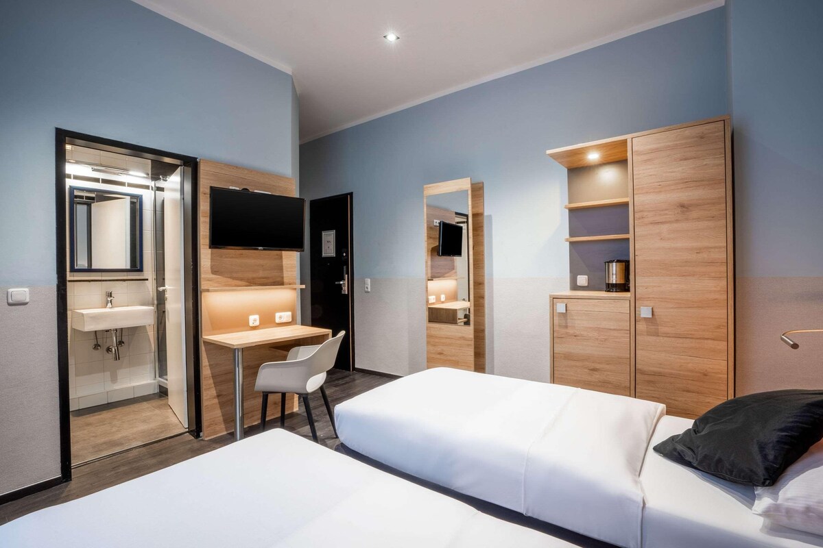 Habitación de hotel con dos camas, un escritorio, una silla, armario, espejo y baño con lavabo y ducha.