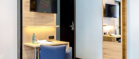Ein modernes Hotelzimmer mit Schreibtisch aus Holz, einem blauen Stuhl, einem Bett mit gemustertem Überwurf, einem an der Wand montierten Flachbildfernseher und einer Tür mit Spiegel.