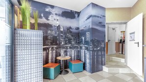 Un moderno vestíbulo de hotel con un mural de la ciudad, un refrigerador, una pequeña mesa redonda y dos taburetes azules.