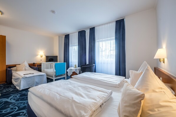 A hotel room with two beds, a desk, a chair, a crib, and a view of the outdoors through large windows. - Hotel Gertrudenhof (Cologne)