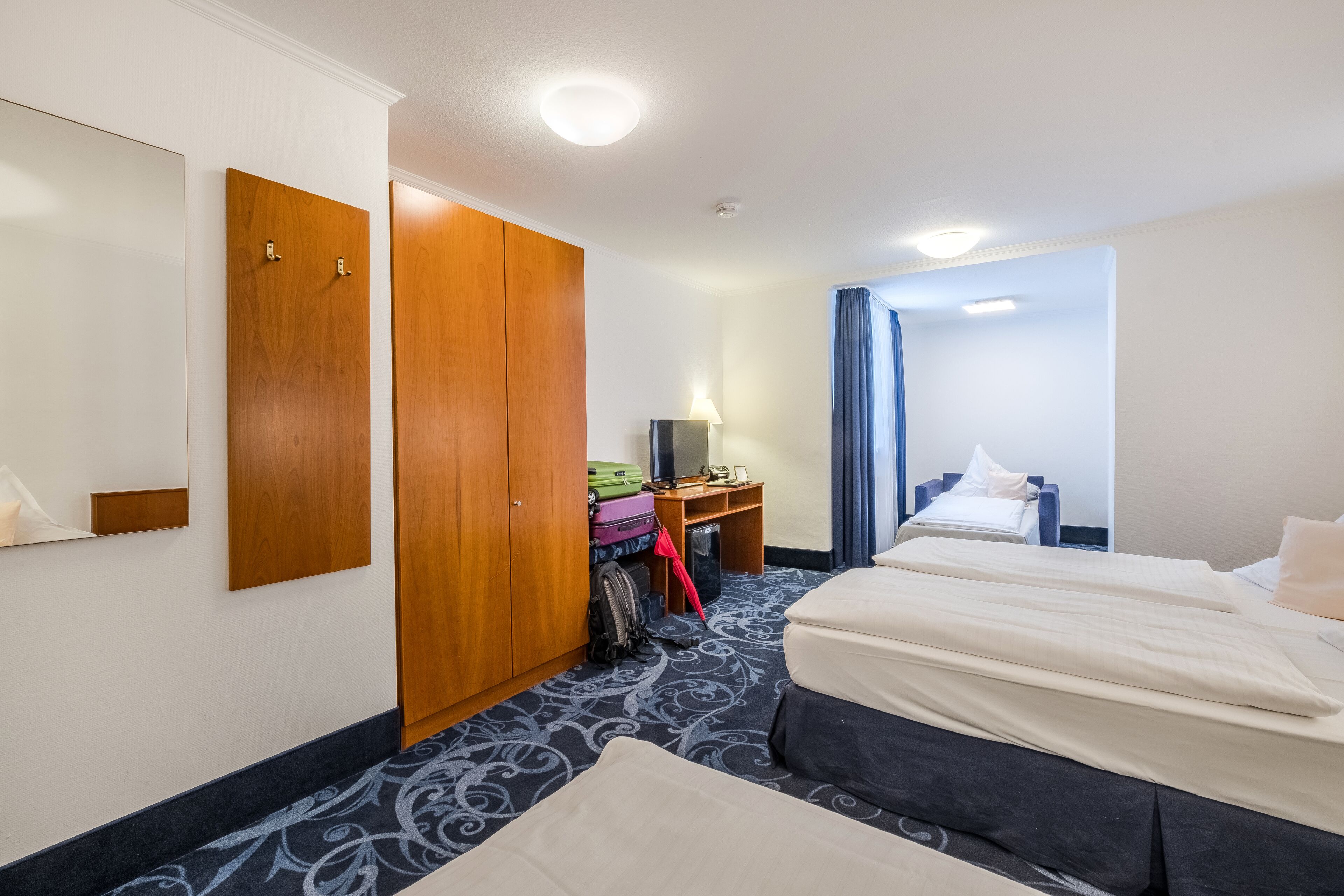 Ein Hotelzimmer mit zwei Betten, einem Kleiderschrank, einem Schreibtisch mit Lampe und einem Koffer.