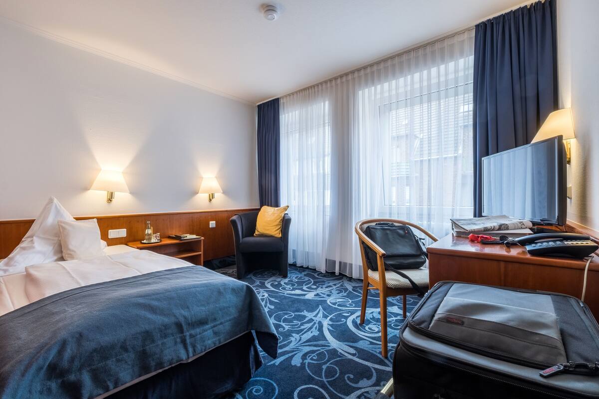 A hotel room with a bed, a desk with a chair, a suitcase, and a window with curtains.