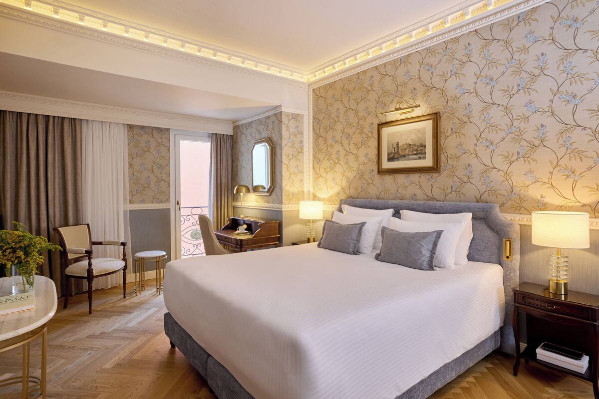 Quarto de hotel com uma cama grande, uma escrivaninha com cadeira, uma mesinha com um vaso e uma janela com cortinas.