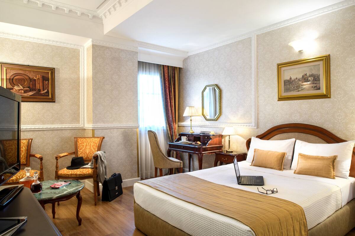Quarto de hotel com cama, secretária com laptop, cadeira, televisão e espelho.