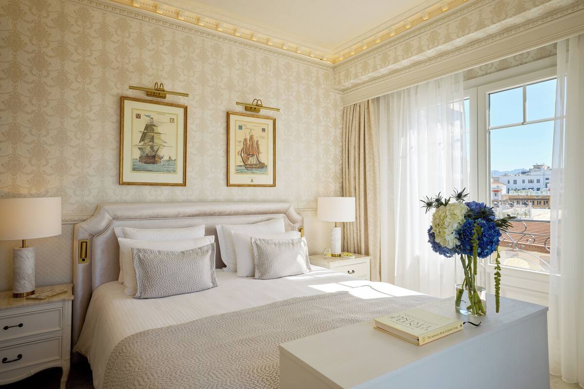 Quarto de hotel com uma cama grande, duas mesas de cabeceira com luminárias, um criado-mudo, um vaso com flores e quadros nas paredes.