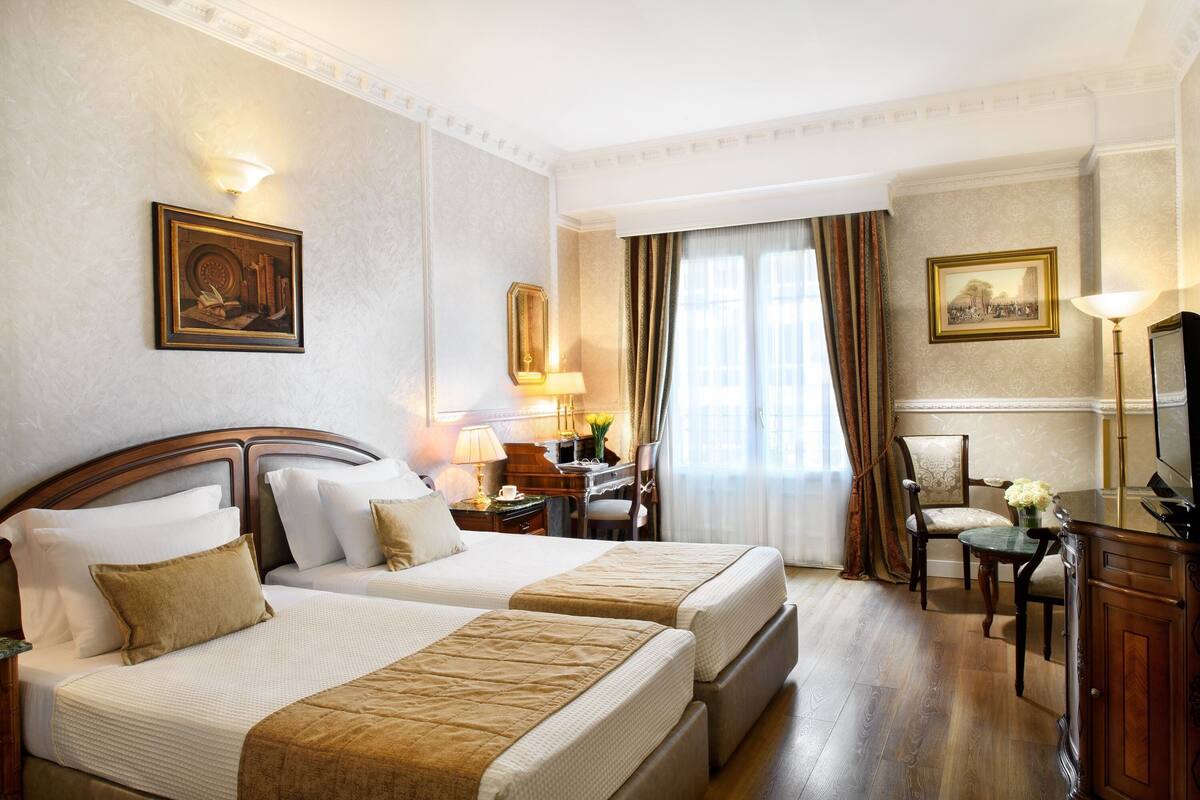 Quarto de hotel com duas camas, uma mesa de trabalho com abajur, uma cadeira e uma televisão.