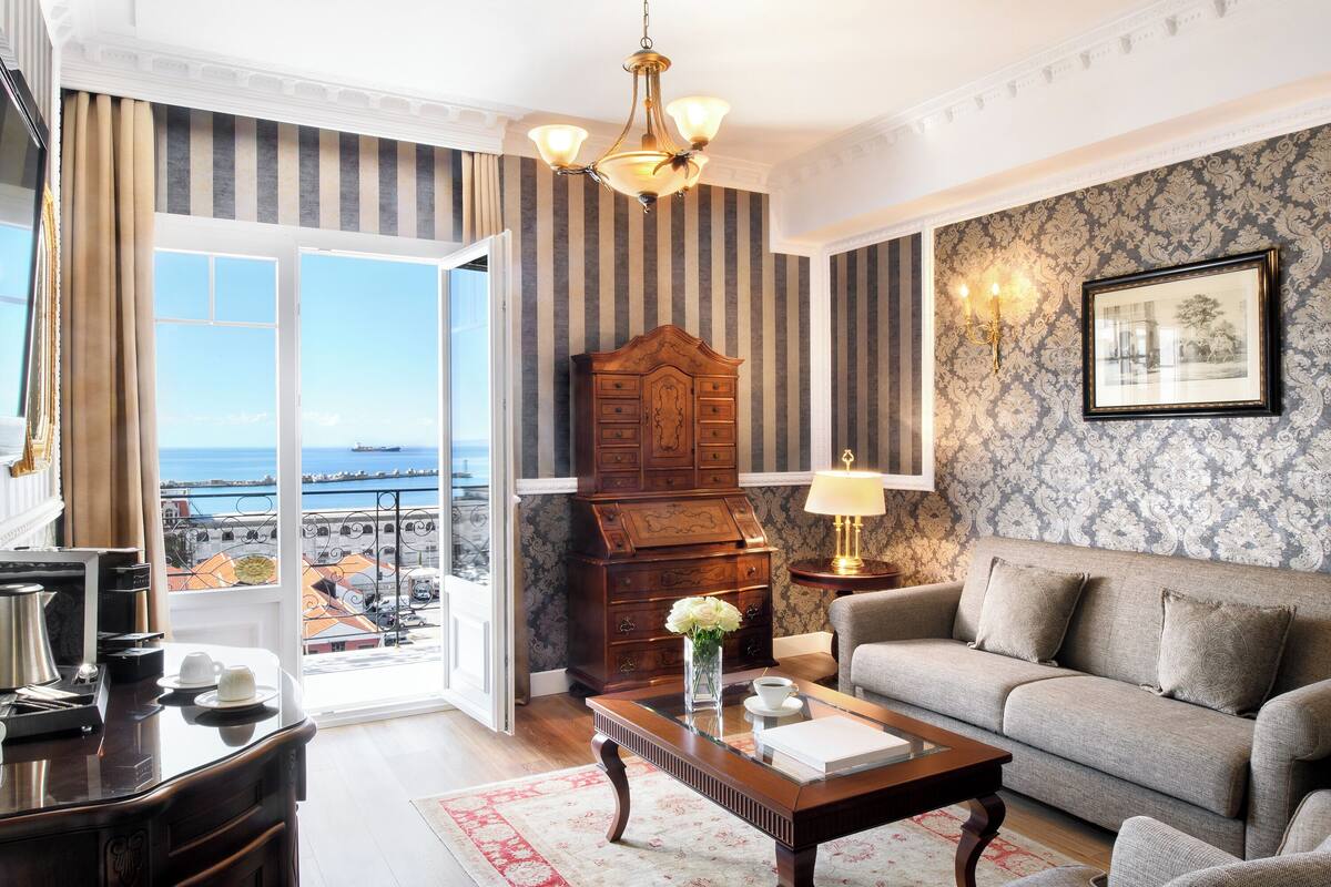 Sala de estar com sofá, armário de madeira, lustre e vista para o mar através de amplas janelas.
