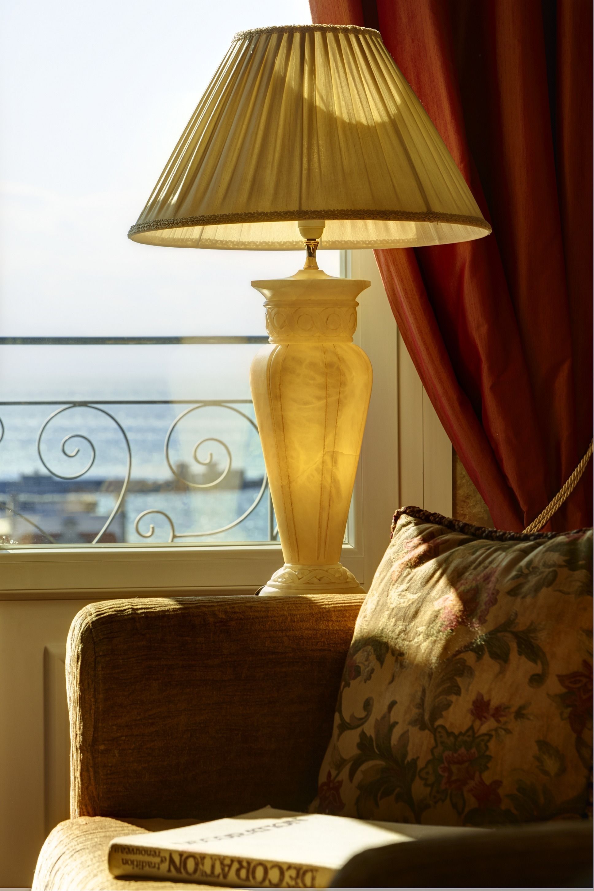 A lamp on a bedside table next to a chair, with a view of the sea through a window with a decorative iron railing.