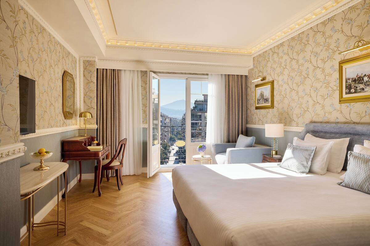 Quarto de hotel com uma cama grande, uma escrivaninha com abajur, uma cadeira e vista da cidade por uma janela ampla.