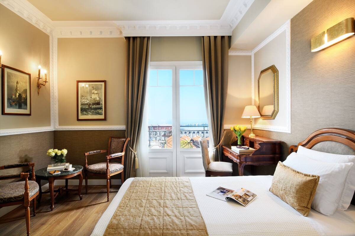 Quarto de hotel com uma cama grande, uma escrivaninha com luminária, duas cadeiras, uma mesinha com um vaso de flores e vista para a cidade pela janela.