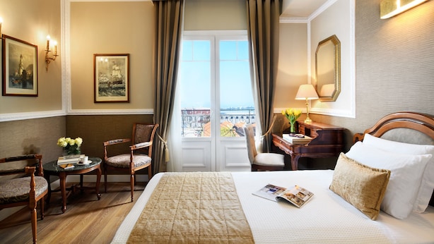 Habitación de hotel con una cama grande, un escritorio con lámpara, dos sillas, una mesa pequeña con un jarrón de flores y vista a la ciudad desde la ventana.