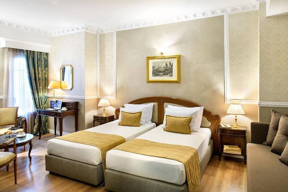Quarto de hotel com duas camas, uma escrivaninha, uma cadeira e um abajur.