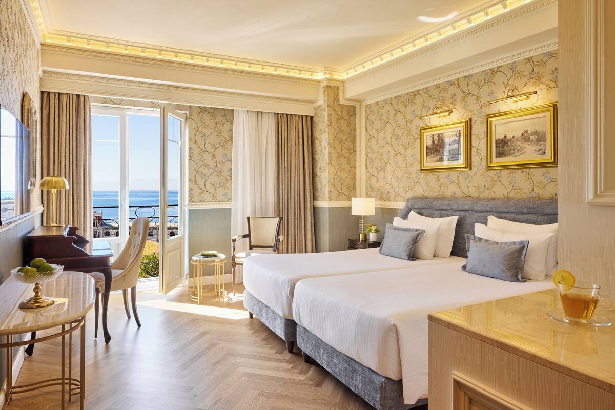 Quarto de hotel com uma cama grande, uma escrivaninha com abajur, uma cadeira e vista para o mar pela janela.