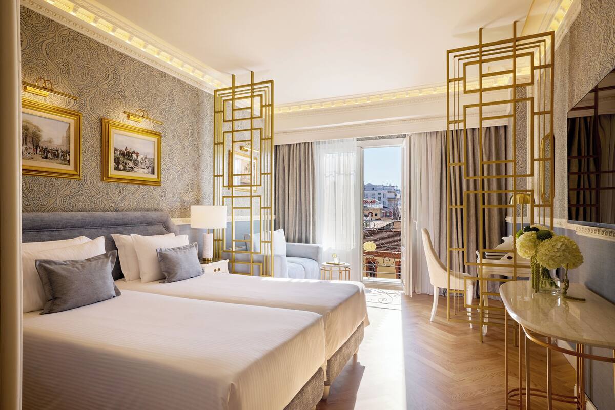 Quarto de hotel com duas camas, varanda, área de refeições e vista da cidade.