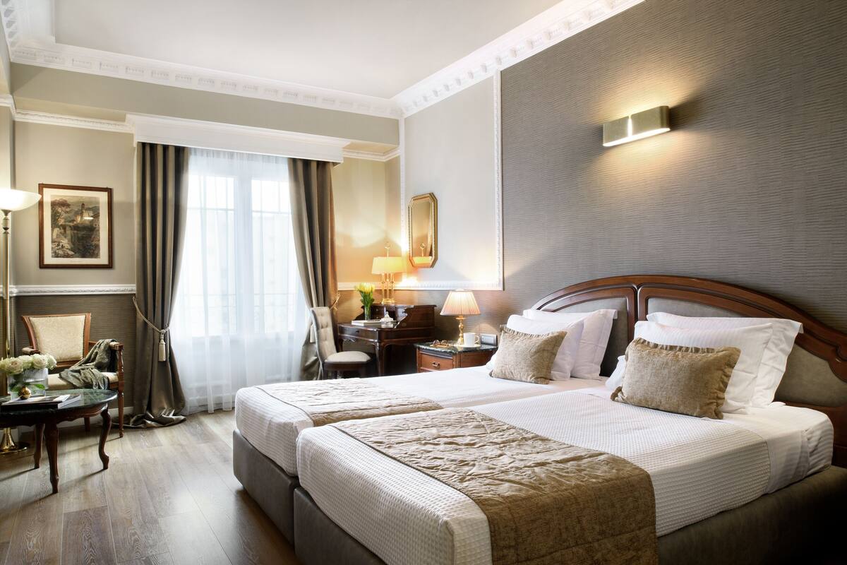 Quarto de hotel com duas camas, uma escrivaninha, uma cadeira e uma janela grande com cortinas.