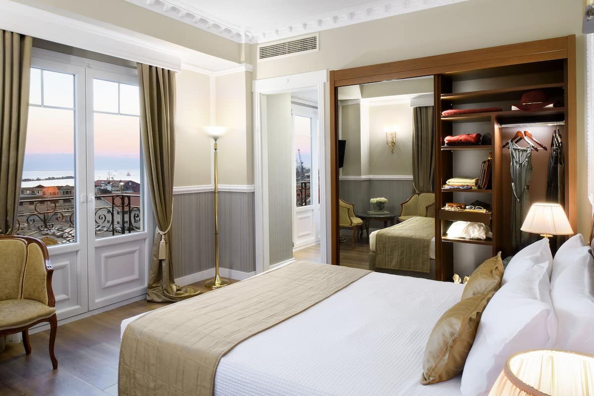 Quarto de hotel com uma cama grande, guarda-roupa, uma cadeira e vista para a cidade através de amplas janelas.