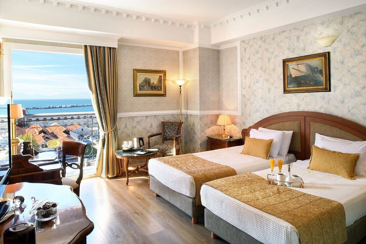 Quarto de hotel com duas camas, uma escrivaninha, uma cadeira, um abajur e vista para a cidade.