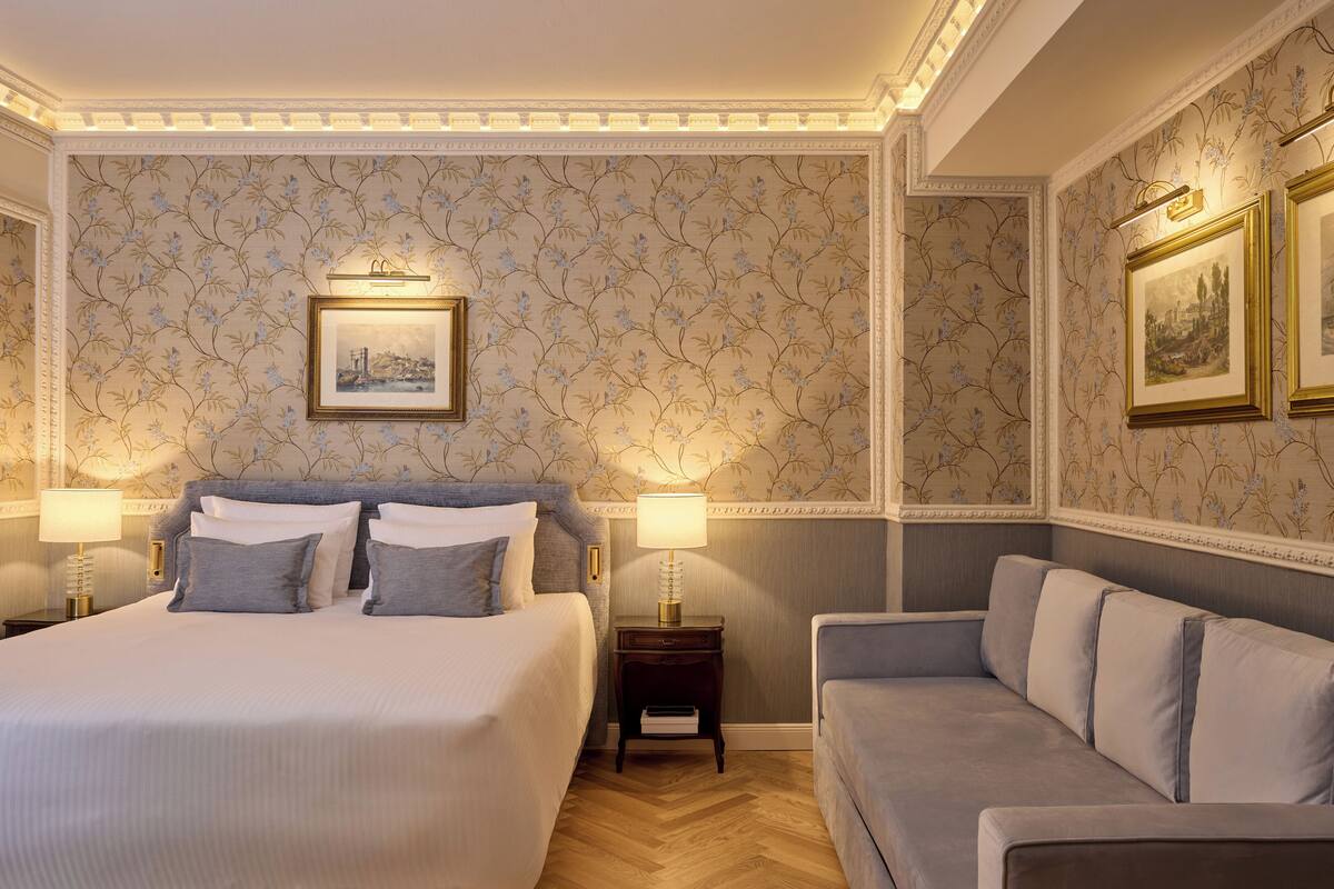 Quarto de hotel com uma cama grande, duas mesas de cabeceira com luminárias, um sofá, papel de parede floral e quadros nas paredes.