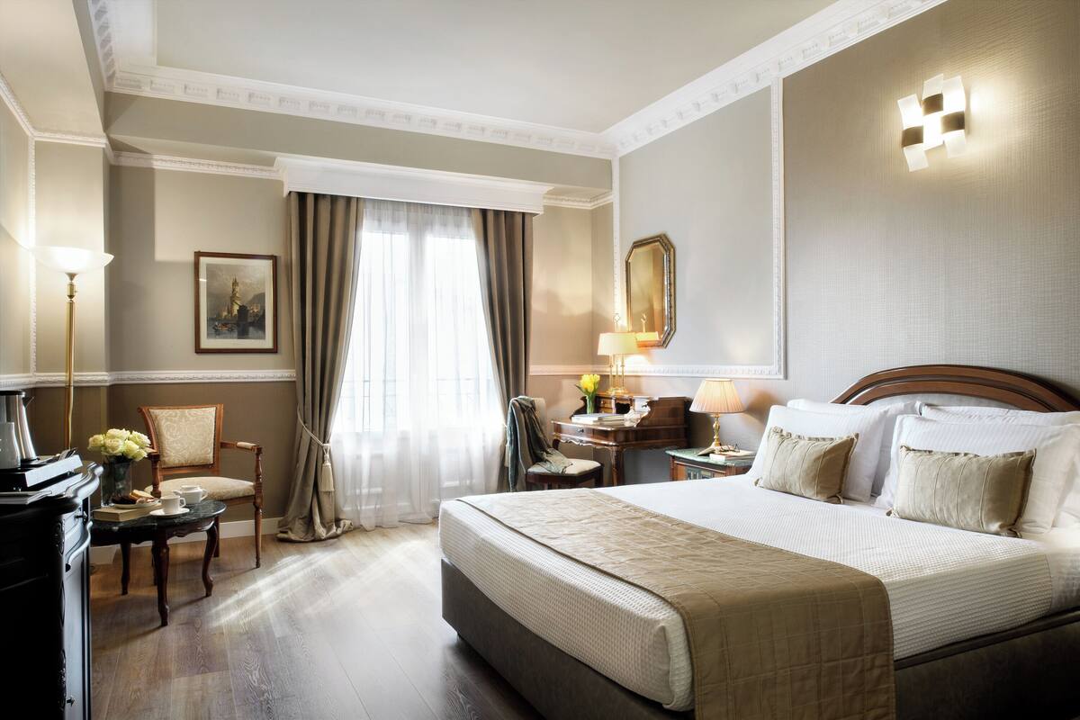 Quarto de hotel com uma cama grande, uma escrivaninha com abajur, uma cadeira e um quadro na parede.