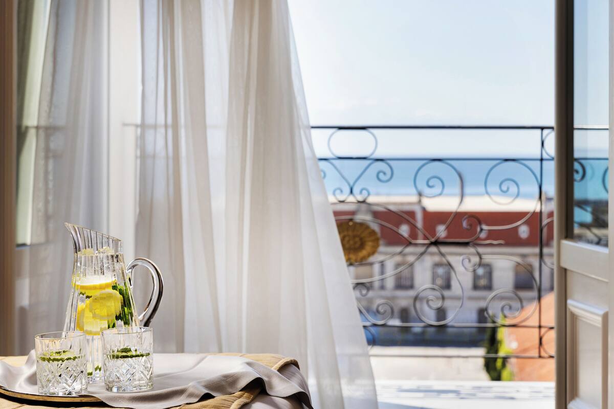 Varanda com grade de ferro forjado, mesa com jarra e copos, e vista de prédios através de cortinas leves.