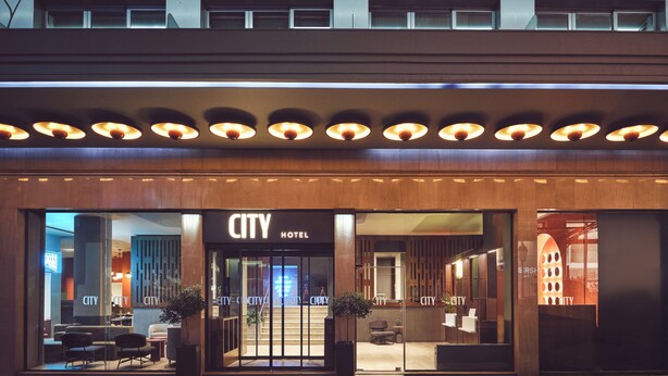 Entrada de un hotel urbano con diseño moderno, que cuenta con fachada de vidrio y un área de estar exterior acogedora.