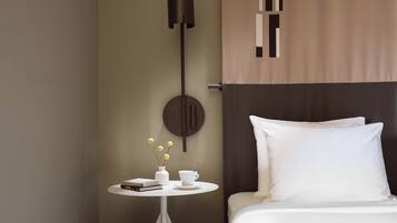 Habitación de hotel ordenada con una cama, una mesita redonda con un florero y libros, una lámpara de pared y una cortina.