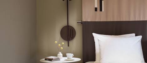 Habitación de hotel ordenada con una cama, una mesita redonda con un florero y libros, una lámpara de pared y una cortina.