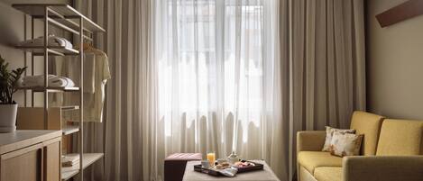 A modern hotel room with a large window, a sofa, a coffee table with breakfast items, and a wardrobe with shelves.