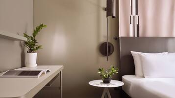 A modern hotel room with a bed, a desk with a laptop, a small round table with a plant, and a wall-mounted light fixture.