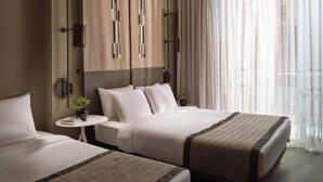 Habitación de hotel con dos camas, una mesita con una planta y un ventanal con cortinas translúcidas.