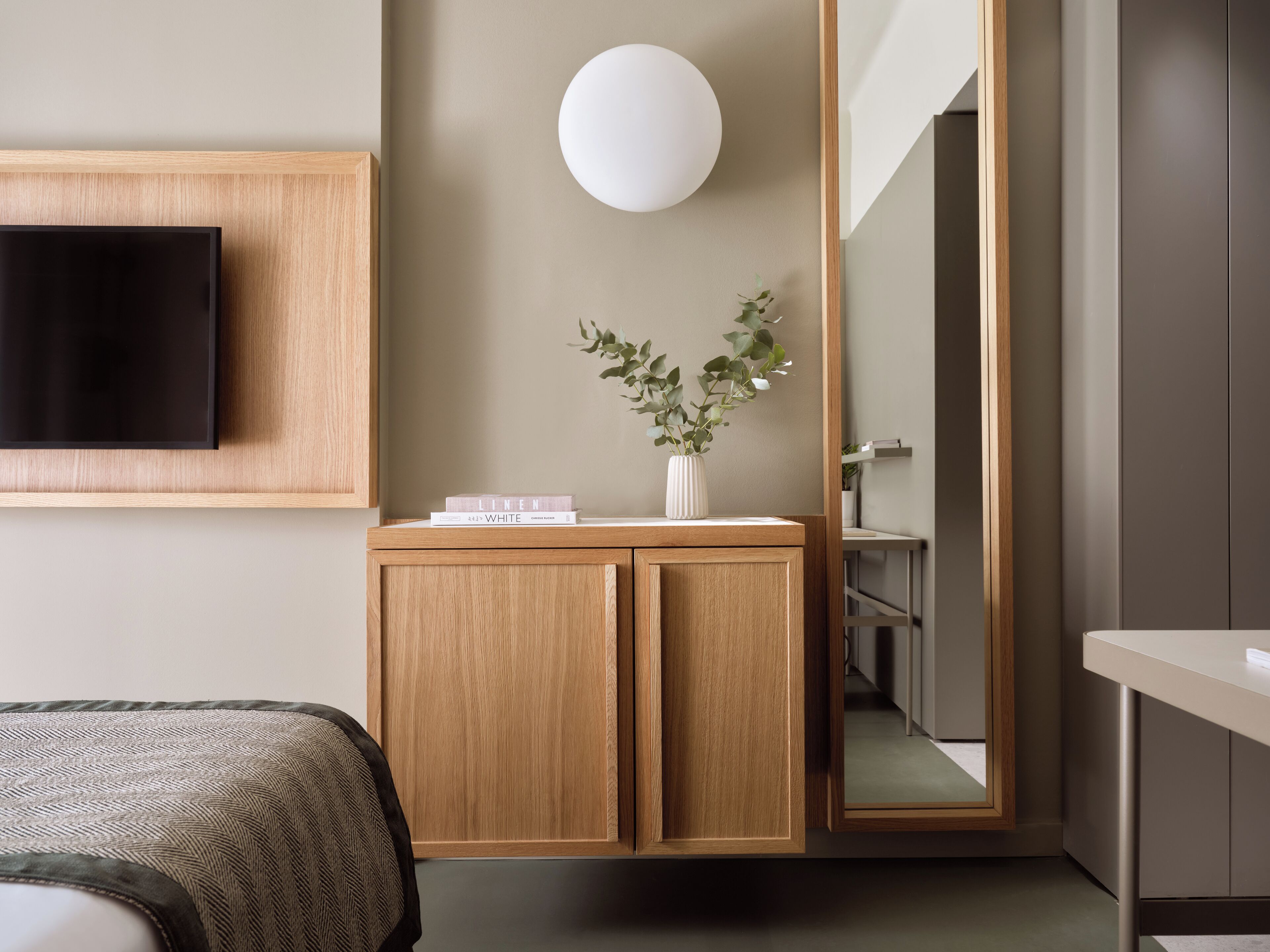 A modern hotel room with a wooden cabinet, a flat-screen TV, a white vase with green leaves, and a round light fixture.