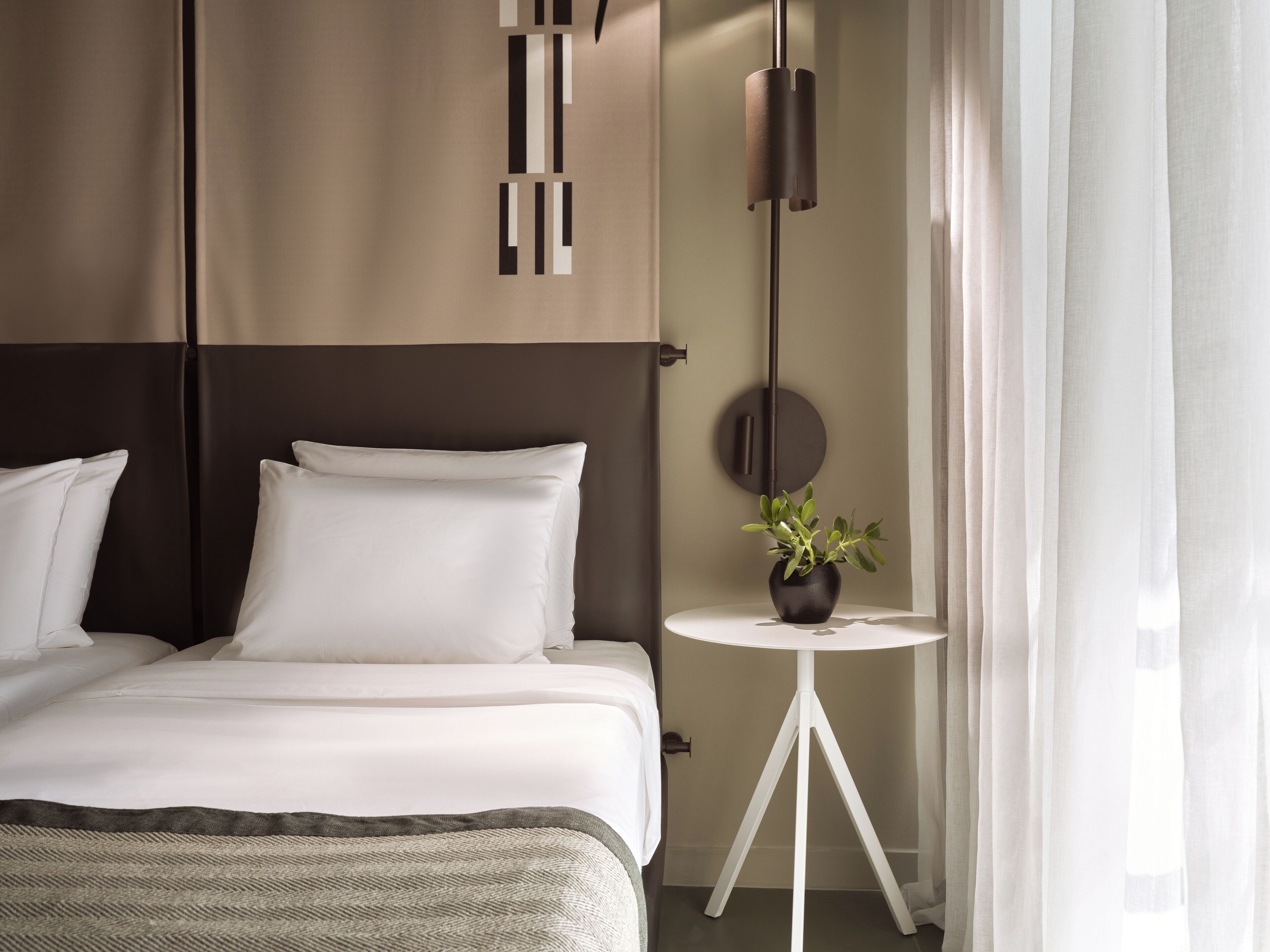 A modern hotel room with a large bed, a bedside table with a potted plant, a round mirror, and a hanging light fixture.