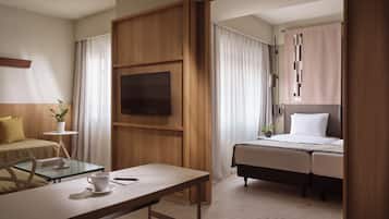 Habitación de hotel con cama, escritorio con una taza y un libro, televisor en la pared y ventana con cortinas.