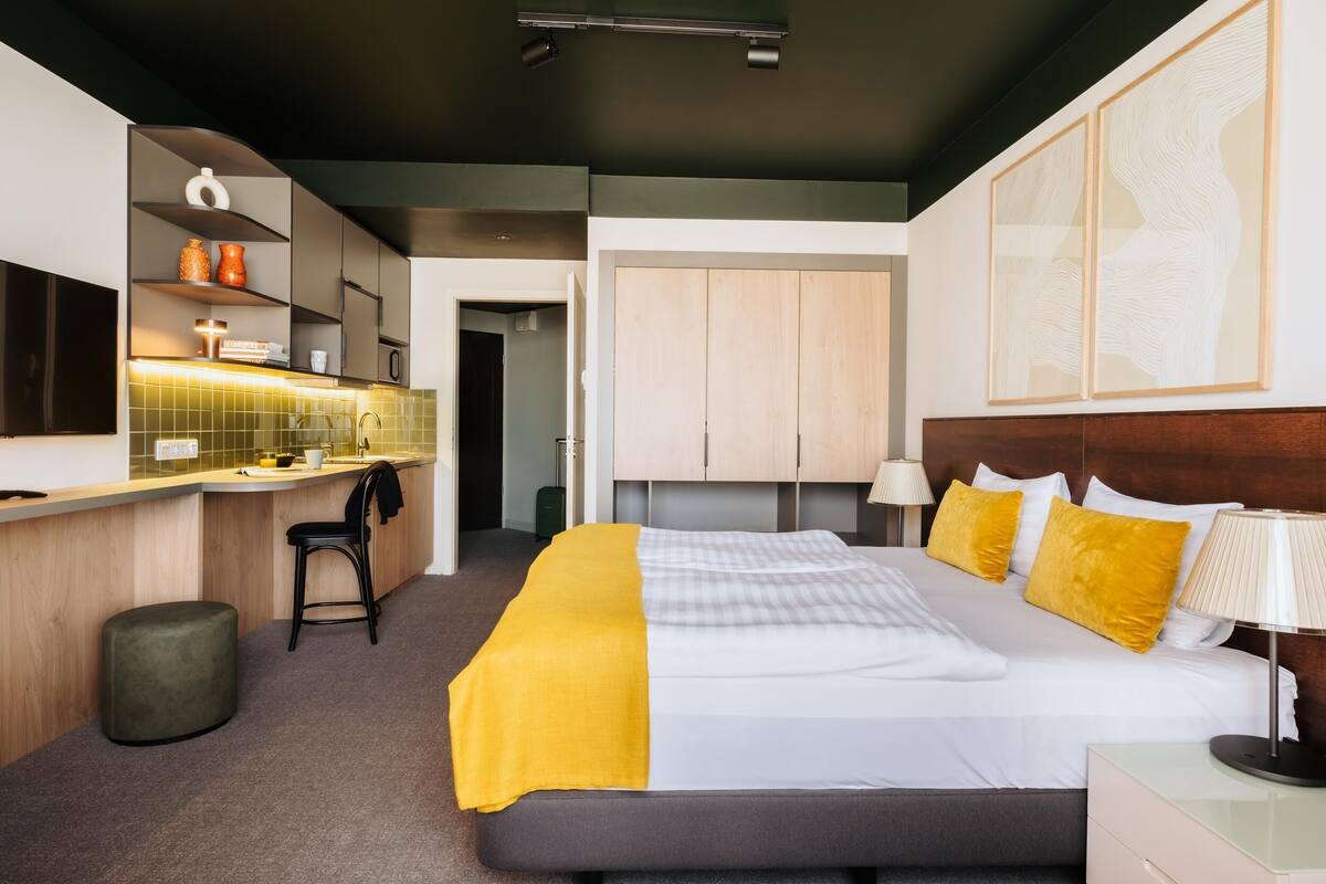 Une chambre d’hôtel moderne équipée d’un lit, d’une table de chevet, d’une lampe et d’un coin cuisine avec une chaise et un comptoir.