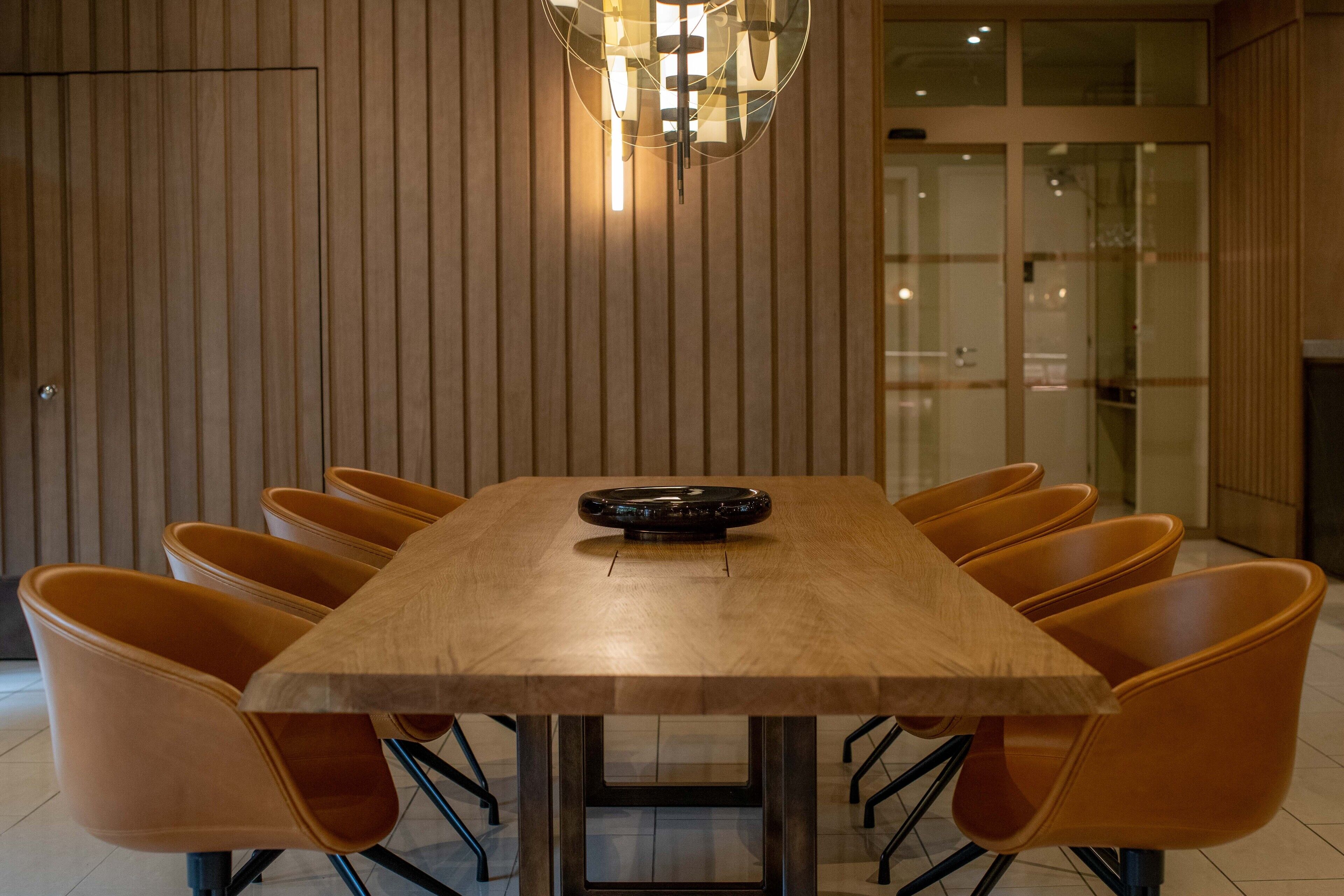 A conference room with a wooden table and orange chairs, a ceiling light fixture, and a glass partition in the background.