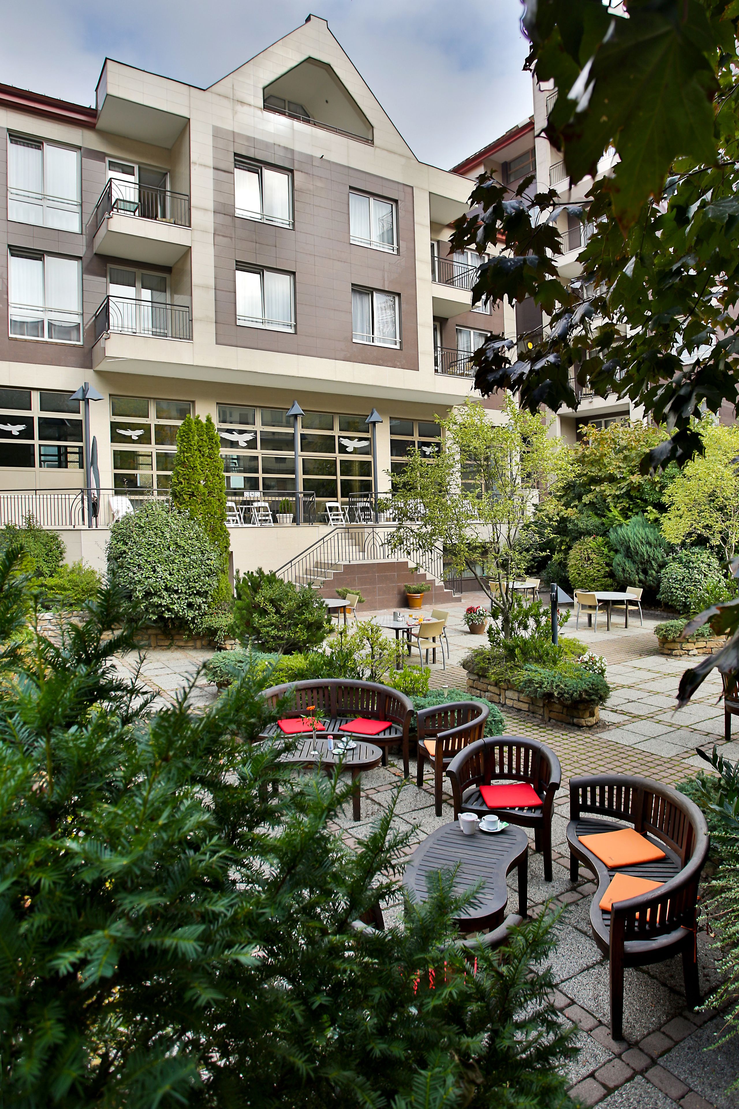 Un patio con mobiliario exterior, rodeado de vegetación y un edificio de varios pisos con balcones.